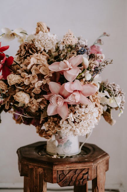 A large floral arrangement featuring a variety of dried and fresh flowers, including pale pink orchids, creamy white hydrangeas, and beige and pink hydrangea blooms, all arranged in a white ceramic va