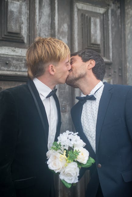 Two men in formal attire, one with blond hair and the other with dark hair, are sharing a kiss in front of a textured gray wooden door. The man with blond hair is holding a bouquet of fresh flowers, i