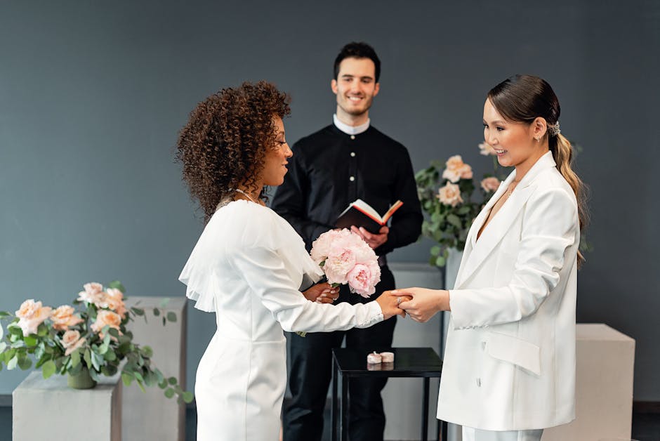 A woman with curly brown hair in a white blazer holding a bouquet of pale pink roses and peonies, standing across from a woman with straight brown hair in a white blazer, smiling and shaking hands in