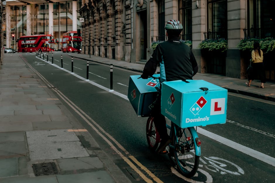 A delivery cyclist riding a blue bicycle alongside a city street, carrying two large blue pizza boxes with Domino's logos attached to the rear and side of the bike. The boxes are secured with straps, 