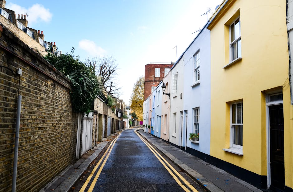 A narrow residential street in Paddington featuring a row of pastel-colored terraced houses painted in soft yellow, pale blue, and white, with white-framed windows and small flower boxes. The street h
